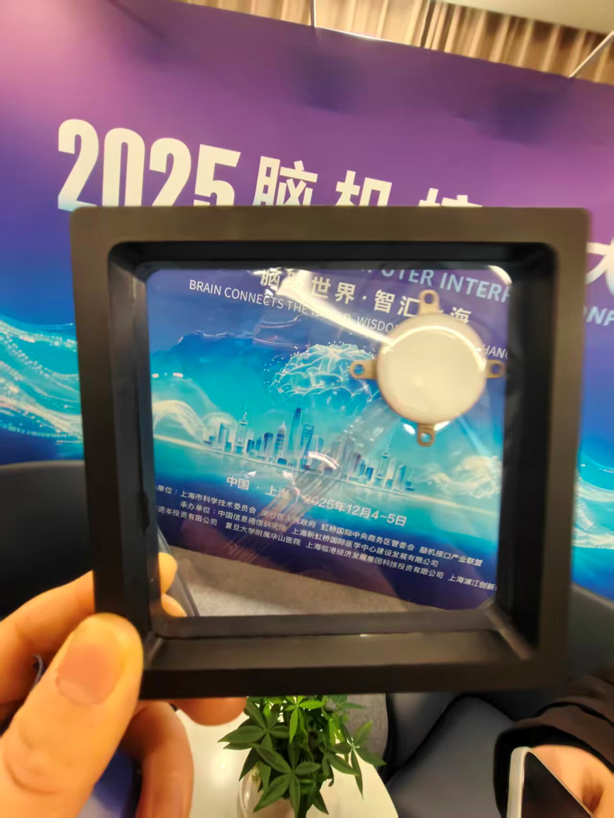 Photograph of the framed WRS02 launch display showing its circular implant body and two fine electrode tails against the 2025 brain-computer-interface conference background.
